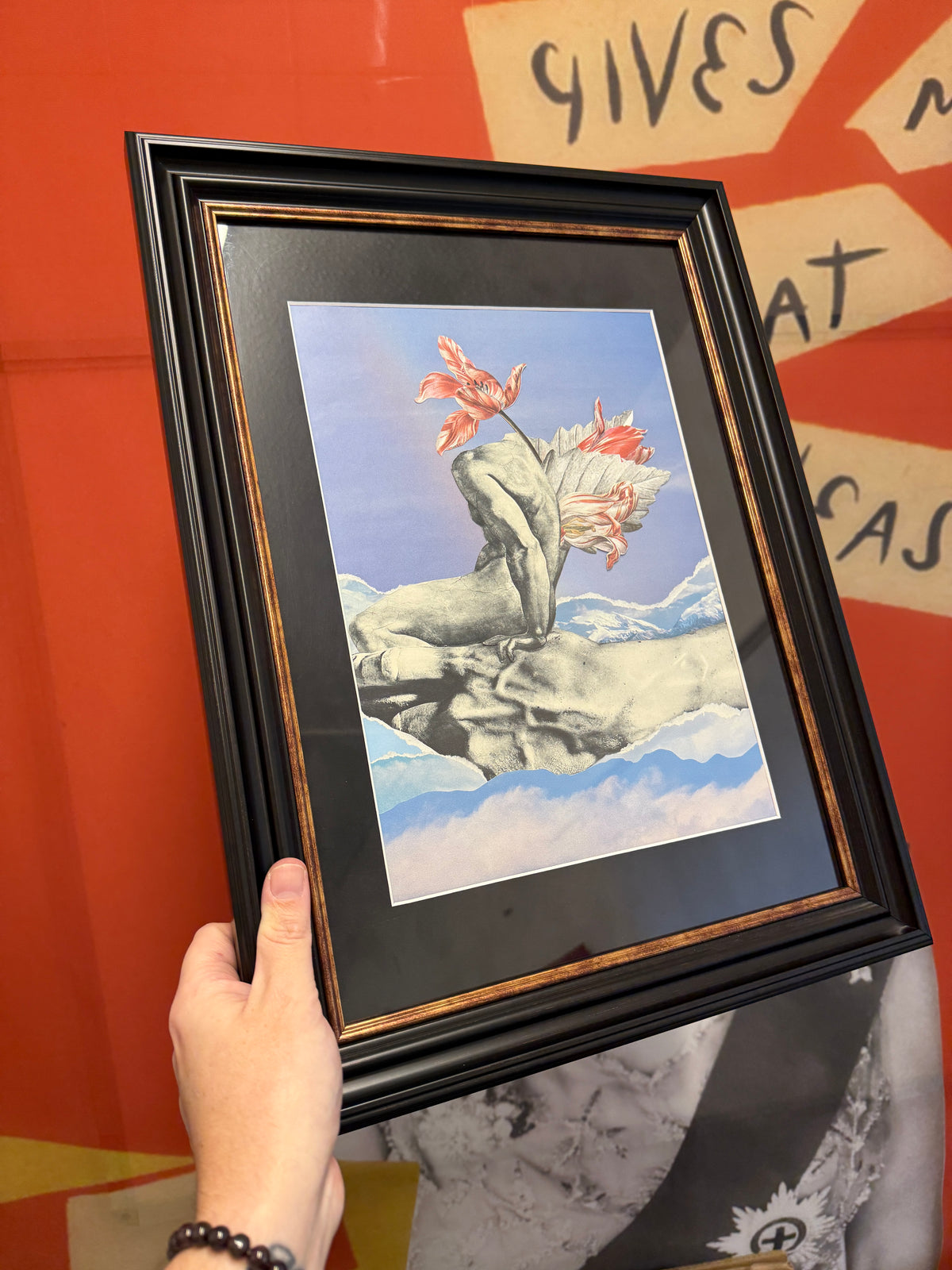 A hand holds a black-framed art print featuring a surreal illustration of a stone statue with red flowers blooming from its head, set against a blue sky and white clouds. The print is matted with a black and gold border, shown against a vibrant orange background with graffiti-style text.